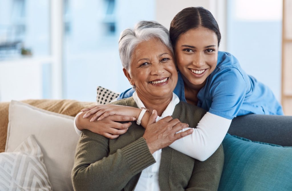 A caregiver hugging an older adult from behind and smiling at the camera in a senior living community's common area.